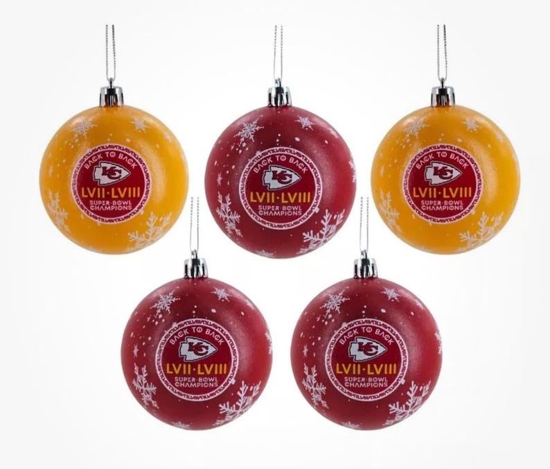 May include: Five Christmas ornaments. Two are yellow, and three are red. Each ornament has the text "BACK TO BACK LVII-LVIII SUPER BOWL CHAMPIONS" in a circular design. White snowflakes are scattered across the ornaments.