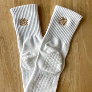 K&ouml;nnte beinhalten: Ein Paar wei&szlig;e Socken mit gerippter Struktur und einer kleinen, gestickten goldenen Blume in der N&auml;he des oberen Randes. Die Sohlen haben ein rutschfestes Muster. Die Socken werden auf einer hellbraunen Holzoberfl&auml;che pr&auml;sentiert.