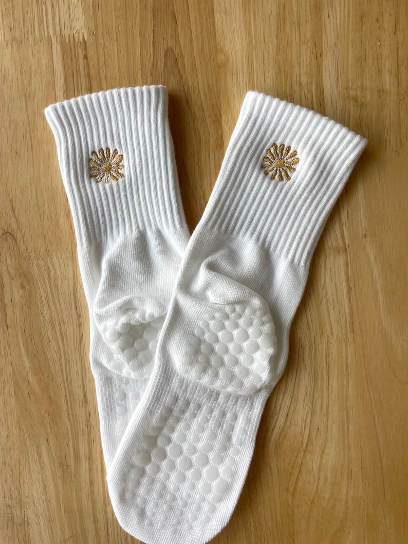 K&ouml;nnte beinhalten: Ein Paar wei&szlig;e Socken mit gerippter Struktur und einer kleinen, gestickten goldenen Blume in der N&auml;he des oberen Randes. Die Sohlen haben ein rutschfestes Muster. Die Socken werden auf einer hellbraunen Holzoberfl&auml;che pr&auml;sentiert.
