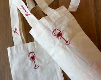 Könnte beinhalten: Zwei cremefarbene Weinflaschentaschen aus Segeltuch mit roter Stickerei eines Weinglases mit herzförmiger Mitte. Die Träger sind mit den Namen Jen und Jonelle in roter Schreibschrift bestickt. Eine Tasche enthält eine Flasche mit Korken.