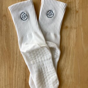 K&ouml;nnte beinhalten: Ein Paar wei&szlig;e Socken mit Rippenmuster und einer dunkelblauen, gestickten Muschel auf jeder Socke. Die Sohlen haben ein rutschfestes Grip-Muster.