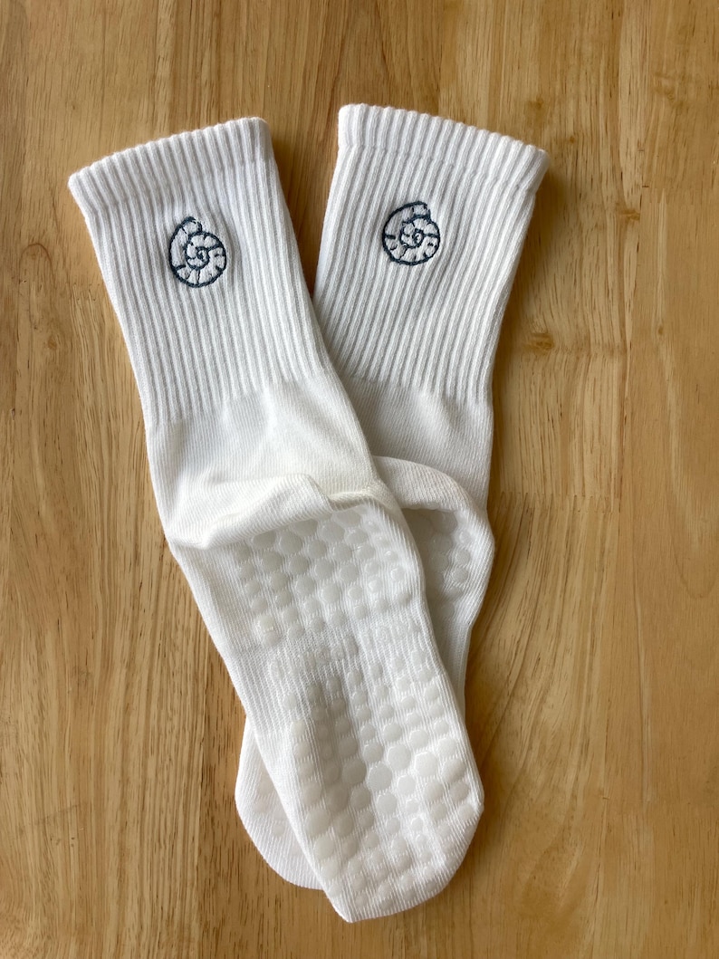 K&ouml;nnte beinhalten: Ein Paar wei&szlig;e Socken mit Rippenmuster und einer dunkelblauen, gestickten Muschel auf jeder Socke. Die Sohlen haben ein rutschfestes Grip-Muster.