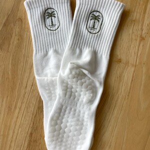 K&ouml;nnte beinhalten: Ein Paar wei&szlig;e Sportsocken mit geripptem B&uuml;ndchen und einem gr&uuml;nen Palmenlogo. Die Sohlen haben ein strukturiertes, rutschfestes Muster. Die Socken sind aus einem weichen, bequemen Material.