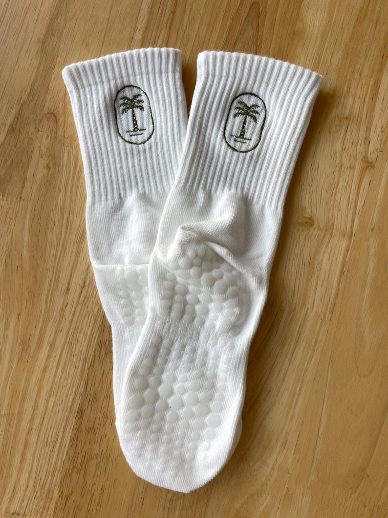K&ouml;nnte beinhalten: Ein Paar wei&szlig;e Sportsocken mit geripptem B&uuml;ndchen und einem gr&uuml;nen Palmenlogo. Die Sohlen haben ein strukturiertes, rutschfestes Muster. Die Socken sind aus einem weichen, bequemen Material.