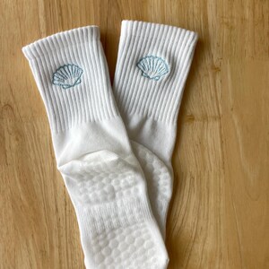 K&ouml;nnte beinhalten: Ein Paar wei&szlig;e gerippte Socken mit hellblauem, gesticktem Muscheldesign am oberen Rand. Die Sohlen haben ein strukturiertes, rutschfestes Muster. Die Socken liegen auf einer hellbraunen Holzoberfl&auml;che.