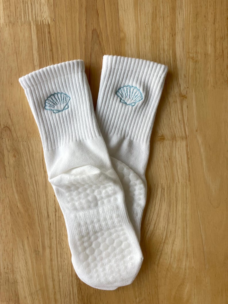 K&ouml;nnte beinhalten: Ein Paar wei&szlig;e gerippte Socken mit hellblauem, gesticktem Muscheldesign am oberen Rand. Die Sohlen haben ein strukturiertes, rutschfestes Muster. Die Socken liegen auf einer hellbraunen Holzoberfl&auml;che.