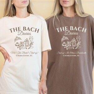 May include: Two t-shirts, one cream and one brown, with the text "THE BACH Diaries" and a graphic of a tiara, perfume, and champagne. The cream shirt says "Me? The Bride? Shut up! Charleston, SC." The brown shirt says "Shut up! It's Olivia's Bachelorette Charleston, SC."