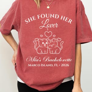 May include: Red-brown t-shirt with white text and graphic. The text reads "SHE FOUND HER Lover" above a heart and two cartoon cats. Below the cats, it says "Mia's Bachelorette" and "MARCO ISLAND, FL &bull; 2026".