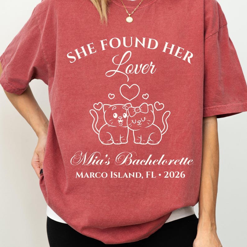 May include: Red-brown t-shirt with white text and graphic. The text reads "SHE FOUND HER Lover" above a heart and two cartoon cats. Below the cats, it says "Mia's Bachelorette" and "MARCO ISLAND, FL &bull; 2026".