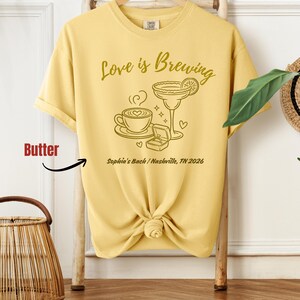 Puede incluir: Camiseta amarilla con la frase "Love is Brewing" y un gr&aacute;fico de una taza de caf&eacute;, un c&oacute;ctel y un anillo de compromiso. El texto "Sophia's Bach / Nashville, TN 2026" tambi&eacute;n est&aacute; impreso.