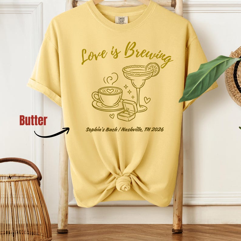 Puede incluir: Camiseta amarilla con la frase "Love is Brewing" y un gr&aacute;fico de una taza de caf&eacute;, un c&oacute;ctel y un anillo de compromiso. El texto "Sophia's Bach / Nashville, TN 2026" tambi&eacute;n est&aacute; impreso.
