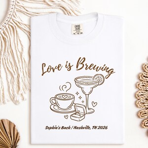 Puede incluir: Camiseta blanca con un dise&ntilde;o marr&oacute;n que dice "Love is Brewing". El dise&ntilde;o incluye una taza de caf&eacute;, una copa de margarita, un joyero con un anillo y el texto "Sophia's Bach / Nashville, TN 2026".