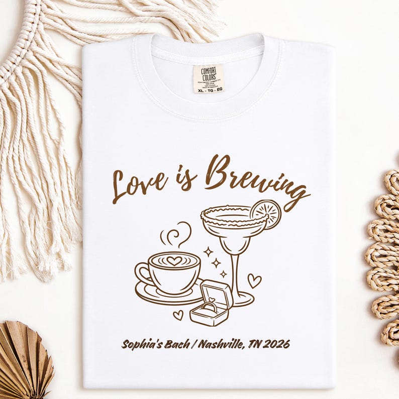 Puede incluir: Camiseta blanca con un dise&ntilde;o marr&oacute;n que dice "Love is Brewing". El dise&ntilde;o incluye una taza de caf&eacute;, una copa de margarita, un joyero con un anillo y el texto "Sophia's Bach / Nashville, TN 2026".