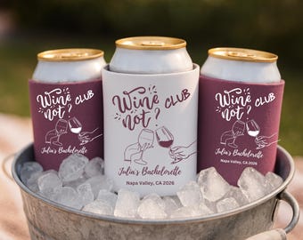 May include: Three beverage can coolers in a metal bucket filled with ice. The coolers are white and burgundy, with the text "Wine Club? Not?" and "Julia's Bachelorette Napa Valley, CA 2026" printed on them. The image has a blurred background.