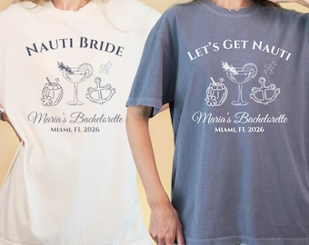 Può includere: Due magliette oversize, una color crema e una blu-grigio, con grafiche a tema nautico. La maglietta color crema recita "NAUTI BRIDE" e la maglietta blu-grigio dice "LET'S GET NAUTI". Entrambe le magliette presentano un'illustrazione di cocktail, ancora e cocco, con "Maria's Bachelorette, Miami, FL 2026".