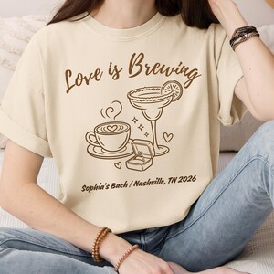 Puede incluir: Camiseta color crema con la frase marr&oacute;n "Love is Brewing". El dise&ntilde;o incluye una taza de caf&eacute;, una copa de margarita, un anillo de compromiso y corazones. Tambi&eacute;n est&aacute; impreso el texto "Sophia's Bach / Nashville, TN 2026".