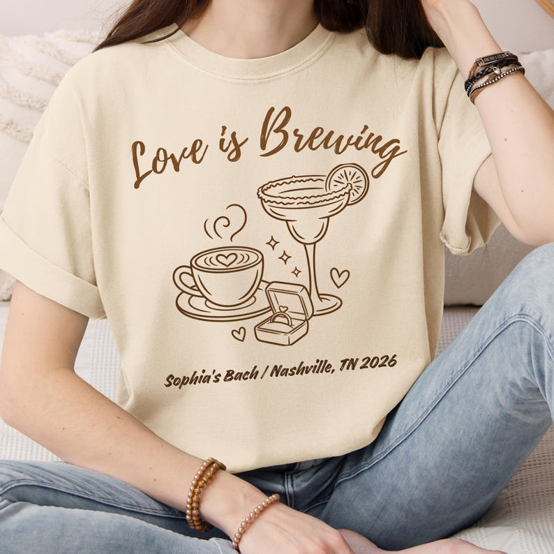 Puede incluir: Camiseta color crema con la frase marr&oacute;n "Love is Brewing". El dise&ntilde;o incluye una taza de caf&eacute;, una copa de margarita, un anillo de compromiso y corazones. Tambi&eacute;n est&aacute; impreso el texto "Sophia's Bach / Nashville, TN 2026".