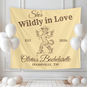 Op de afbeelding: Een beige achtergrond voor een vrijgezellenfeest met de tekst "She's Wildly in Love" en "Olivia's Bachelorette Nashville, TN". Een cartoon cheetah met een cowboyhoed houdt cocktails vast. Witte ballonnen staan op de voorgrond.