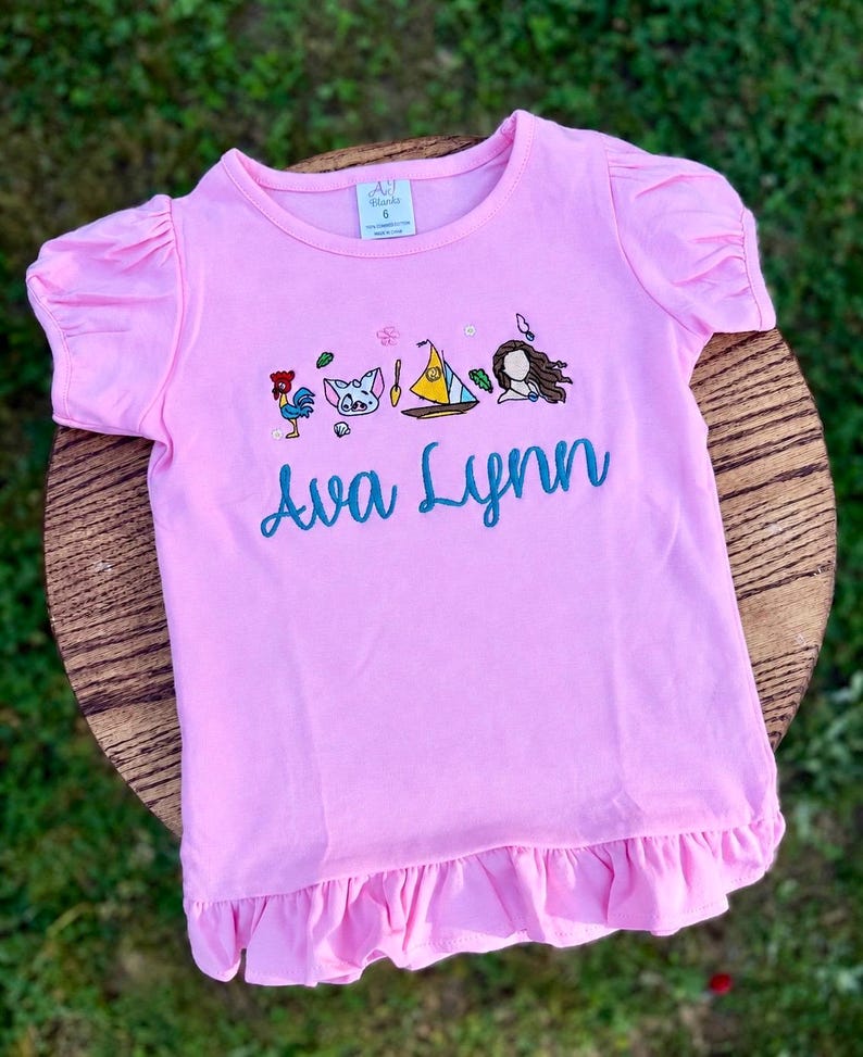 May include: A light pink short-sleeved t-shirt with ruffled sleeves and a ruffled hem. The shirt features an embroidered design of a rooster, a pig, a sailboat, and a person, with the name Ava Lynn embroidered in teal script below the icons.