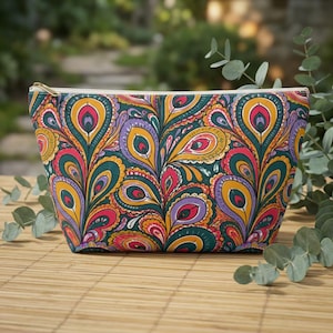 May include: A zipped pouch featuring a vibrant, repeating peacock feather pattern in shades of teal, mustard yellow, magenta, and purple. The bag has a tapered, trapezoidal shape with a gold-toned metal zip closure along the top edge.