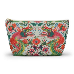 May include: A rectangular cosmetic bag with a colorful dragon and floral pattern. The design features two dragons facing each other, with red, green, and yellow details, set against a light background. The bag has a gold zipper.