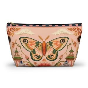 May include: A zippered pouch with a light pink background featuring a large central moth with orange, green, and cream wings. The design includes two green pagodas, stylized clouds, orange foliage, and a dark border with pink dots at the top.