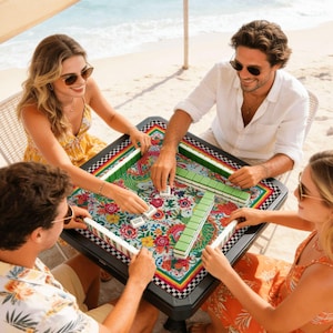 May include: A group of four people playing Mahjong on a black square table. The table has a colorful floral and dragon design. The players are seated around the table on a beach, with the ocean in the background.