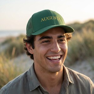 May include: A green baseball cap with the word "AUGUSTA" embroidered in gold. The cap has a mesh back and a curved brim. The person in the image is wearing a green button-down shirt.