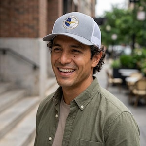 May include: A light gray and white trucker hat featuring a circular patch on the front. The patch displays a blue and white design with a gold bird emblem and the text SANTA MARIA ACORES in a circular border. The hat has a structured front panel.
