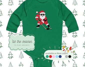 Puede incluir: Un body de bebé verde de manga larga con un gráfico de Santa Claus con gafas de sol jugando al hockey sobre hielo. El texto dice 'tis the season TO PLAY HOCKEY!'. Un pequeño gráfico muestra los colores disponibles: verde, rojo, rosa, azul, azul marino, blanco, gris y negro.