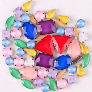 Puede incluir: Una colección de gemas facetadas de colores en varias formas, incluyendo redondas, de lágrima y rectangulares. Las gemas son en tonos de azul, morado, rojo, rosa, amarillo y verde, con engastes de metal plateado. Ideal para la fabricación de joyas o adornos.