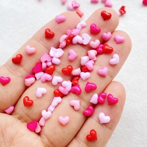 May include: A handful of small, heart-shaped embellishments in shades of red, pink, and light pink. These decorative hearts are scattered across the palm, creating a vibrant and romantic display. Perfect for crafts or adding a touch of love.