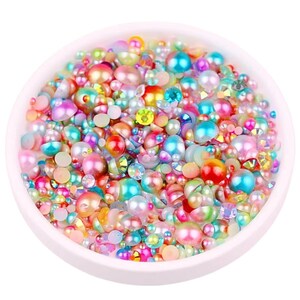 Può includere: Una ciotola bianca piena di un assortimento di perline e gemme colorate e iridescenti. Le perline variano per dimensioni e forma, tra cui rotonde, piatte e sfaccettate, con colori come rosa, blu, verde, giallo e arancione. La disposizione è densa e visivamente accattivante.