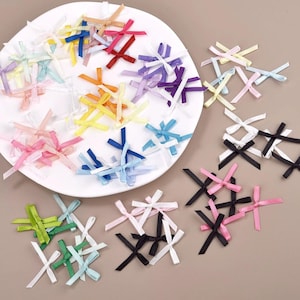 Puede incluir: Una colección de lazos de cinta de raso de colores en varios tonos, incluyendo rosa, azul, amarillo y morado. Algunos lazos están colocados en un plato blanco, mientras que otros están dispersos sobre una superficie neutra. Ideales para manualidades y adornos.