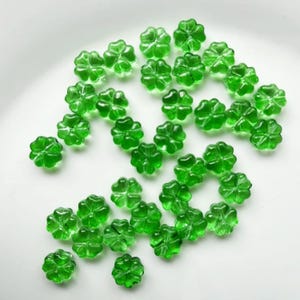 May include: A collection of translucent green four-leaf clover beads. The beads are scattered across a white surface, showcasing their detailed clover shape and vibrant color. Ideal for crafting or jewelry making.