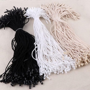 May include: Three bundles of hang tag fasteners in black, white, and beige. Each fastener consists of a thin cord with a small plastic locking mechanism at the end. The bundles are arranged side by side, displaying the varied colors of the cords.