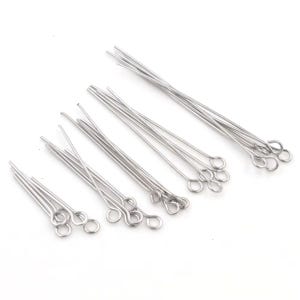 May include: A collection of silver-colored eye pins, arranged on a white surface. The pins have a loop at one end and a straight wire extending from the loop. The eye pins are used for jewelry making and crafting.