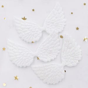 May include: Four white, glittery angel wing embellishments are arranged on a white background with gold star and dot confetti. The wings have detailed feather patterns and are likely for crafts or decorations.
