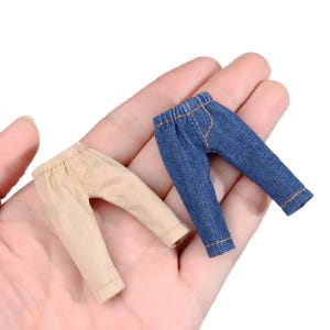 以下が含まれることがあります： 2組のミニチュア人形用パンツ。1組は薄いタン色、もう1組は青色のデニムです。どちらのペアも、伸縮性のあるウエストバンドとステッチのディテールを備えた、シンプルでクラシックなデザインです。パンツは小さく、人形やその他の小さなフィギュア用です。