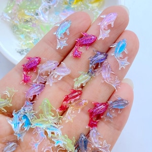 Puede incluir: Una colección de pequeños dijes iridiscentes con forma de pez en varios colores, incluyendo rosa, azul y verde. Los dijes están esparcidos sobre una mano, mostrando su calidad translúcida y sus intrincados detalles. Estos dijes decorativos son ideales para proyectos de manualidades.