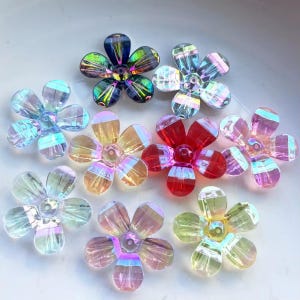 Puede incluir: Una colección de cuentas iridiscentes en forma de flor en varios colores, incluyendo verde, azul, rojo, amarillo y rosa. Cada cuenta tiene cinco pétalos y una cuenta central. Las cuentas están hechas de un material translúcido.