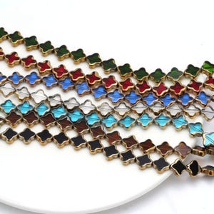 May include: Rows of clover-shaped beads in various colors, including green, red, blue, clear, brown, turquoise, and black. Each bead has a gold-colored border. The beads are arranged on a white surface, suitable for jewelry making.