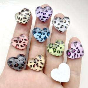 May include: A collection of heart-shaped embellishments in various colors, including pink, blue, and yellow, each adorned with a leopard print pattern. The embellishments are made of a clear, glossy material.
