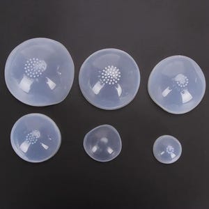 Könnte beinhalten: Eine Sammlung von sechs transparenten Silikon-Brusthütchen in verschiedenen Größen. Jedes Hütchen ist kreisförmig mit einem zentralen Bereich aus kleinen Perforationen. Die Hütchen sind auf einer dunklen Oberfläche angeordnet.