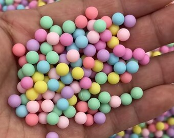 Puede incluir: Un puñado de pequeñas cuentas redondas y coloridas en tonos pastel de rosa, azul, verde, amarillo y morado. Las cuentas son lisas y de color uniforme, creando una textura lúdica y visualmente atractiva. Probablemente se utilizan para manualidades o fines decorativos.