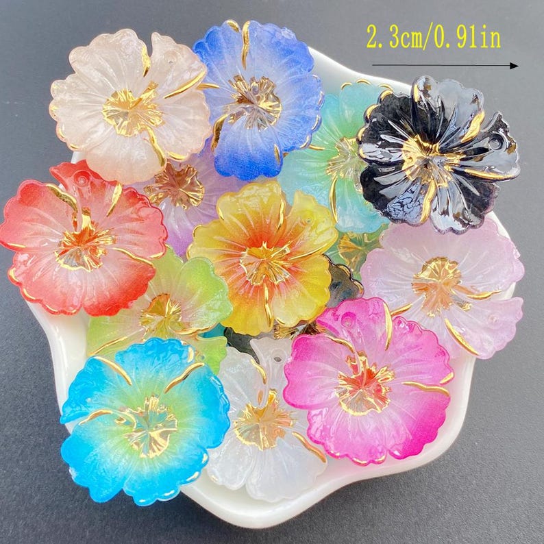 May include: A collection of colorful flower-shaped embellishments, each approximately 0.91 inches (2.3 cm) in diameter. The flowers feature a variety of colors, including pink, blue, yellow, and white, with gold-colored accents. They are arranged in a white dish.