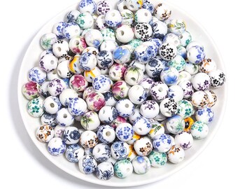 Può includere: Una collezione di perline in ceramica bianca con motivi floreali in vari colori, tra cui blu, rosa e verde, disposte su un piatto bianco. Le perline rotonde hanno un foro al centro, adatte per la creazione di gioielli o lavori artigianali.