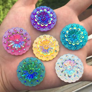 以下が含まれることがあります： 7つのカラフルな、丸い、花の形をした装飾品のコレクション。各ピースは、中央に花の模様があり、キラキラとしたアクセントとスカラップエッジが特徴です。色はピンク、紫、黄色、青、白など。クラフトに最適です。