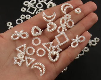 May include: A collection of white pearl embellishments in various shapes, including hearts, moons, squares, and bows. The assortment is displayed against a dark background, showcasing the delicate details of each piece. These decorative elements are ideal for craft projects.