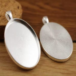 Peut inclure: Deux supports de pendentif ovales argentés avec des boucles pour attacher une chaîne. L'un est vide, montrant l'intérieur, l'autre a un dos texturé. Les supports sont sur une surface en bois.
